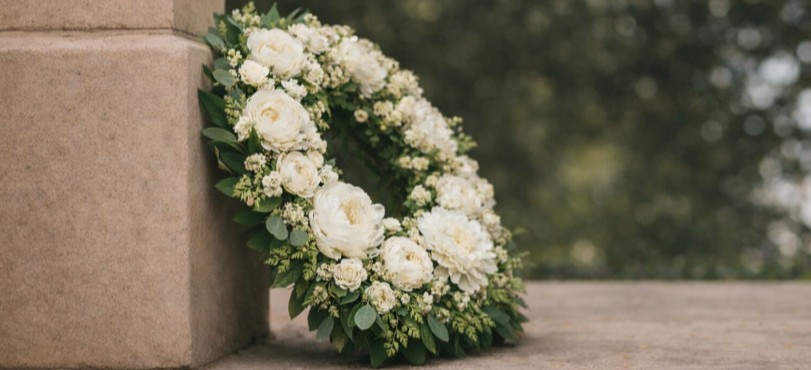 En vakker krans med ulike blomster og grønne blader, plassert ved en steinmur. De jordnære tonene og detaljene i blomstene gir et naturlig og verdig uttrykk, med en uskarp bakgrunn som skaper dybde i bildet. Foto.
