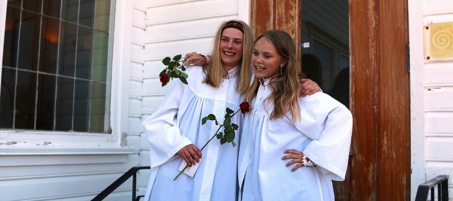 En gruppe ungdommer i hvite kapper står smilende og klare til konfirmasjon. De ser glade og forventningsfulle ut, med en lys og moderne bakgrunn som fremhever høytidsstemningen. Foto.