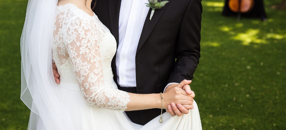 Et symbolsk bilde av et nygift par som holder hender, der begge bærer gifteringer. Bruden er kledd i en elegant, hvit kjole, mens brudgommen har på seg en klassisk, mørk dress. Bildet fokuserer på hendene og formidler en følelse av kjærlighet og samhold. Foto.